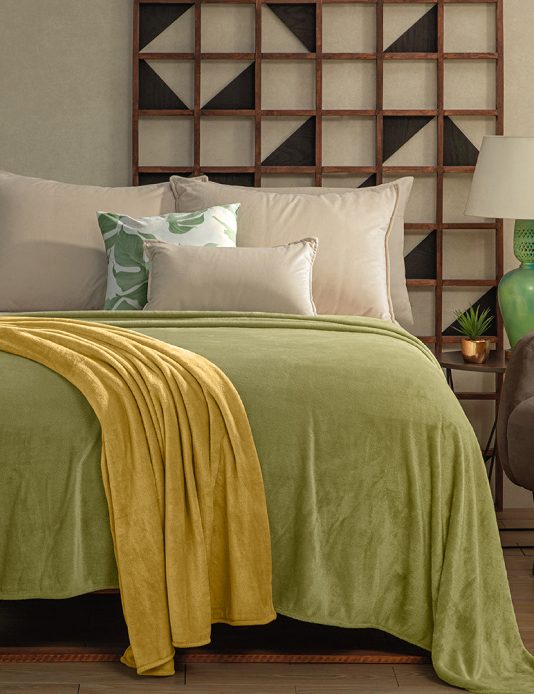 A neatly made bed with the Cobertor Flannel Ligero Ontario Verde by Intima Hogar, beige and green pillows, geometric wooden wall art above, and a bedside table with a lamp and small plant for lasting comfort.