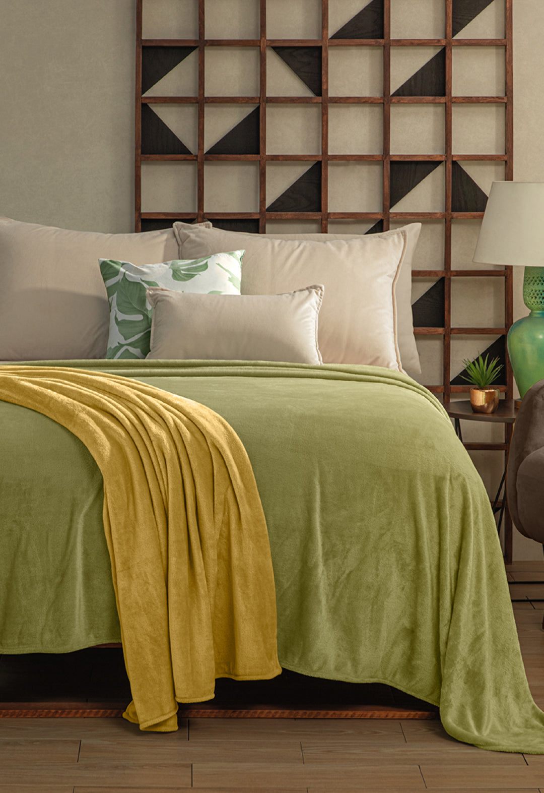 A neatly made bed with the Cobertor Flannel Ligero Ontario Verde by Intima Hogar, beige and green pillows, geometric wooden wall art above, and a bedside table with a lamp and small plant for lasting comfort.