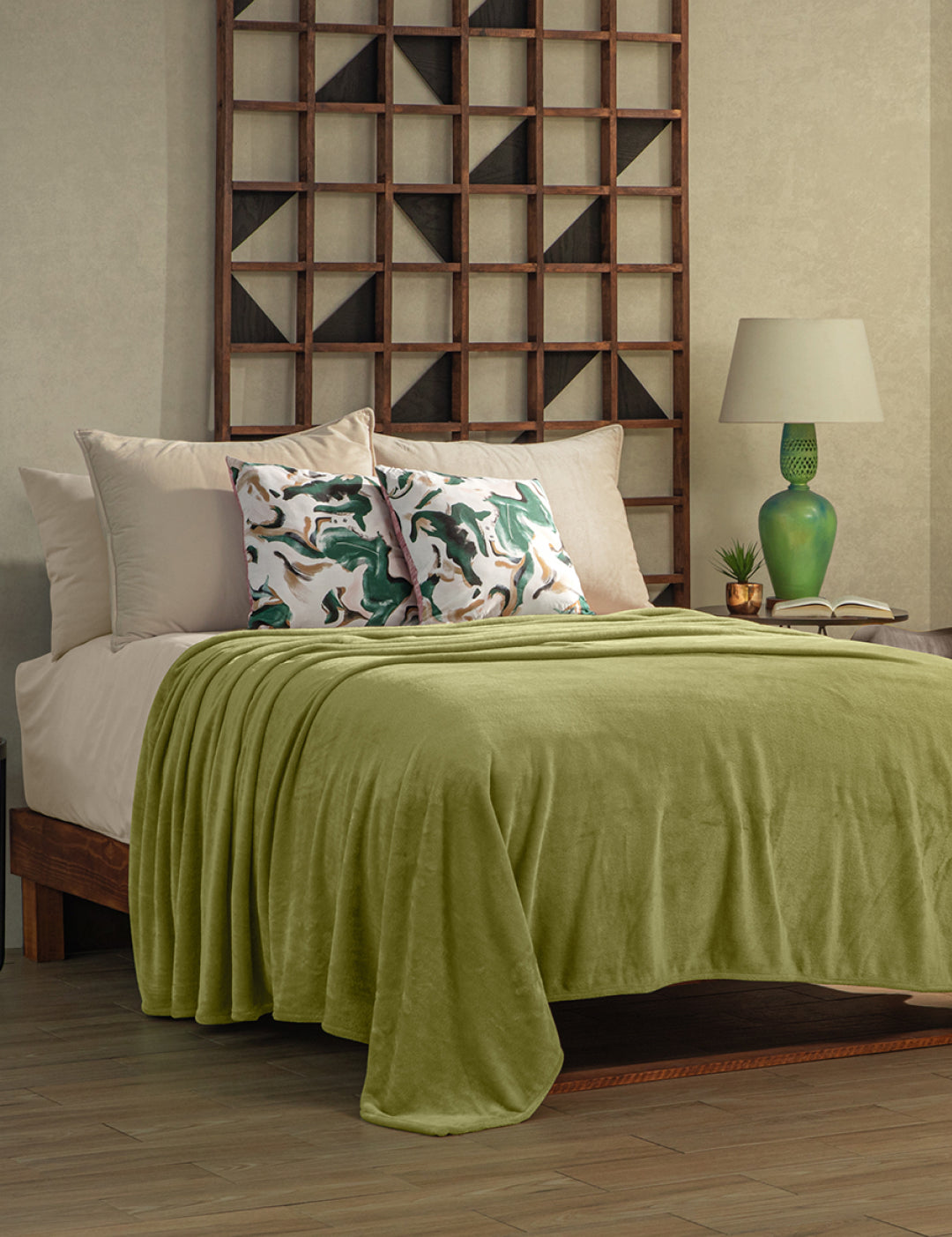 A neatly made bed with the Intima Hogar Cobertor Flannel Ligero Ontario Verde, beige sheets, and two pillows with green and white abstract designs. A wooden shelf, lamp, and green vase add lasting comfort to the modern room.