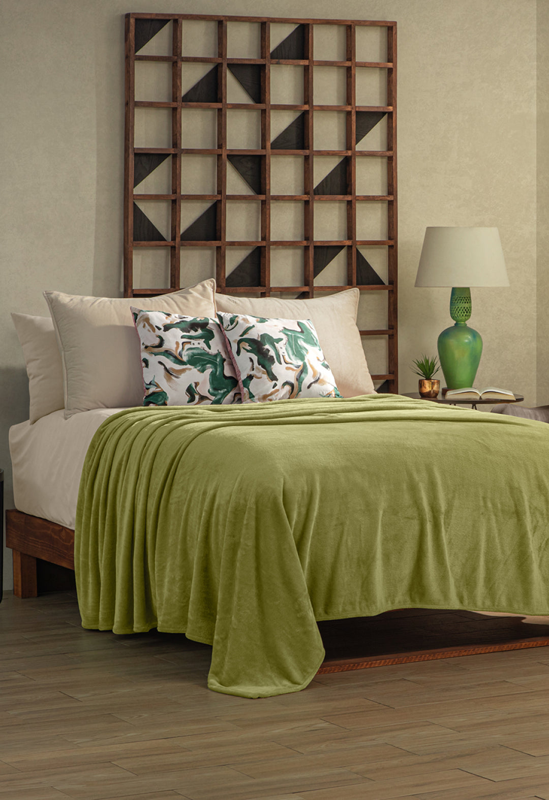 A neatly made bed with the Intima Hogar Cobertor Flannel Ligero Ontario Verde, beige sheets, and two pillows with green and white abstract designs. A wooden shelf, lamp, and green vase add lasting comfort to the modern room.