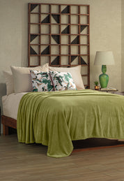 A neatly made bed with the Intima Hogar Cobertor Flannel Ligero Ontario Verde, beige sheets, and two pillows with green and white abstract designs. A wooden shelf, lamp, and green vase add lasting comfort to the modern room.