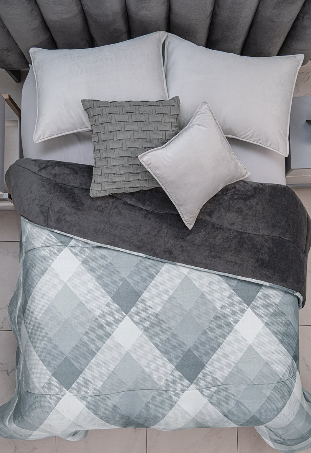 A neatly made bed features the Cobertor Flannel Extra Suave Palacio by Intima Hogar in gray and white checks, a dark gray throw, and four pillows—three light gray, one with a woven pattern—on a soft, upholstered headboard.