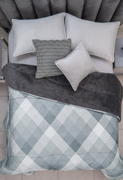 A neatly made bed features the Cobertor Flannel Extra Suave Palacio by Intima Hogar in gray and white checks, a dark gray throw, and four pillows—three light gray, one with a woven pattern—on a soft, upholstered headboard.