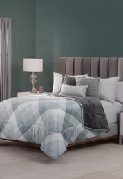 A modern bedroom with a tall gray upholstered headboard, layered gray and white bedding topped with the Cobertor Flannel Extra Suave Palacio by Intima Hogar, decorative pillows, a bedside table with lamp, and a dark green accent wall.