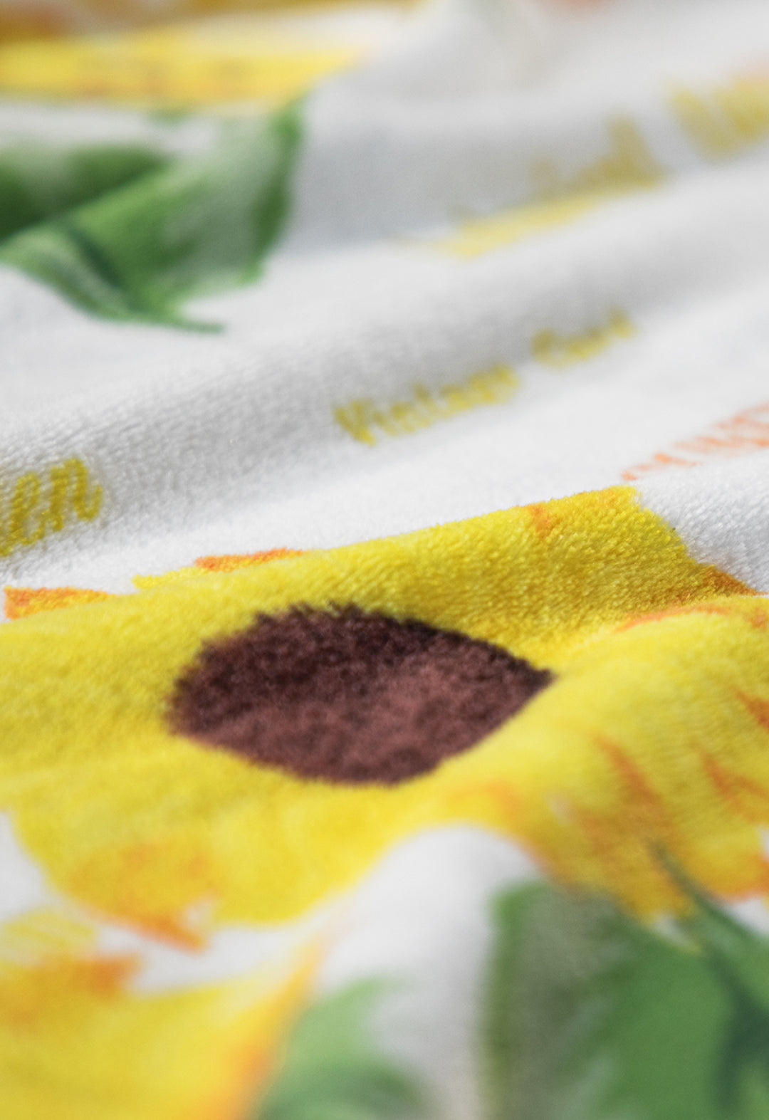 Close-up of Cobertor Flannel Ligero Sun by Intima Hogar: soft flannel blanket with a sunflower print—bright yellow flower, dark brown center, green leaves on white. Botanical design, resistente al lavado.