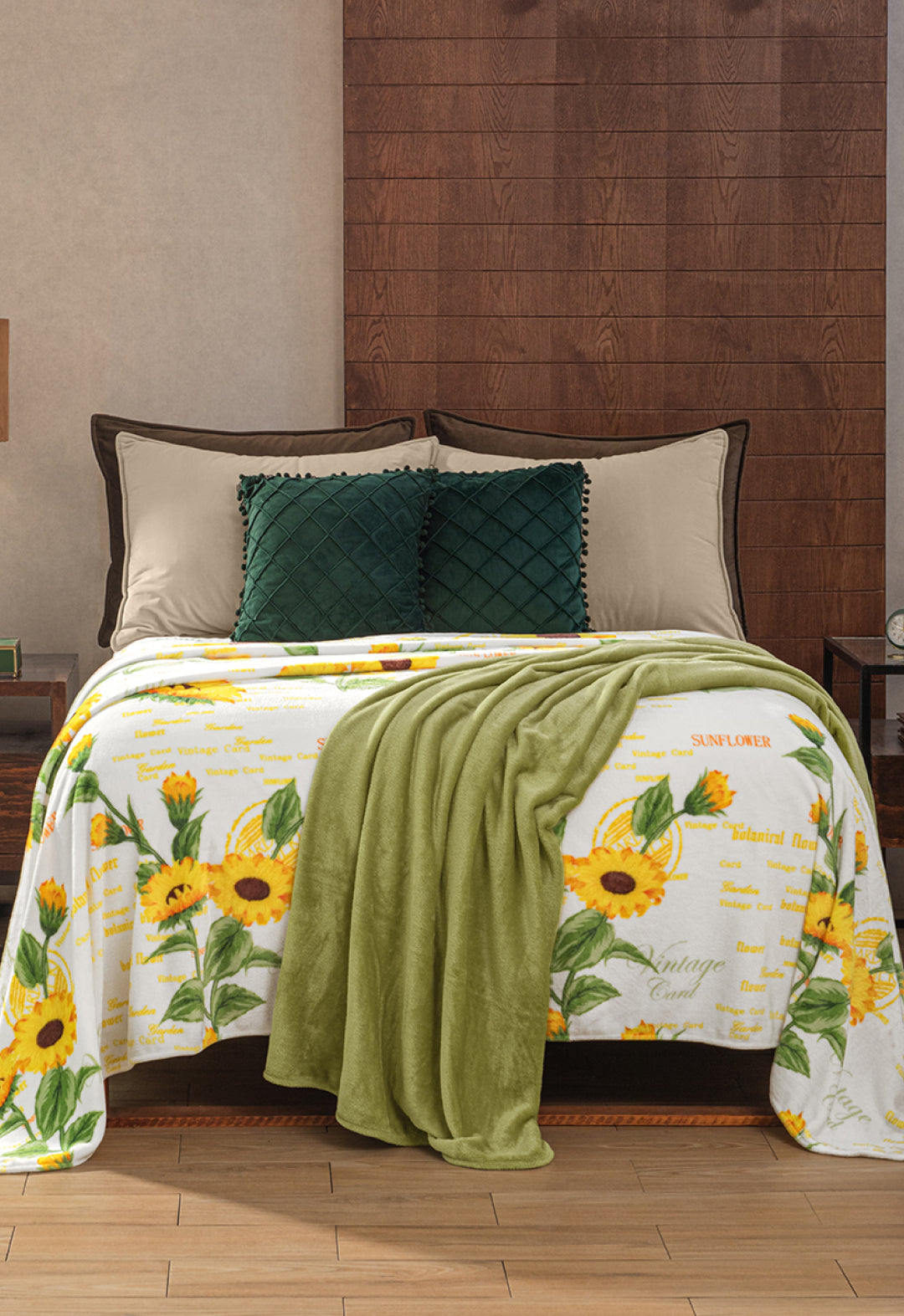 A bed with cream sheets, two dark green pillows, and the Cobertor Flannel Ligero Sun by Intima Hogar, featuring a sunflower diseño botánico and text. A green throw blanket is at the foot, and the headboard is dark wood.