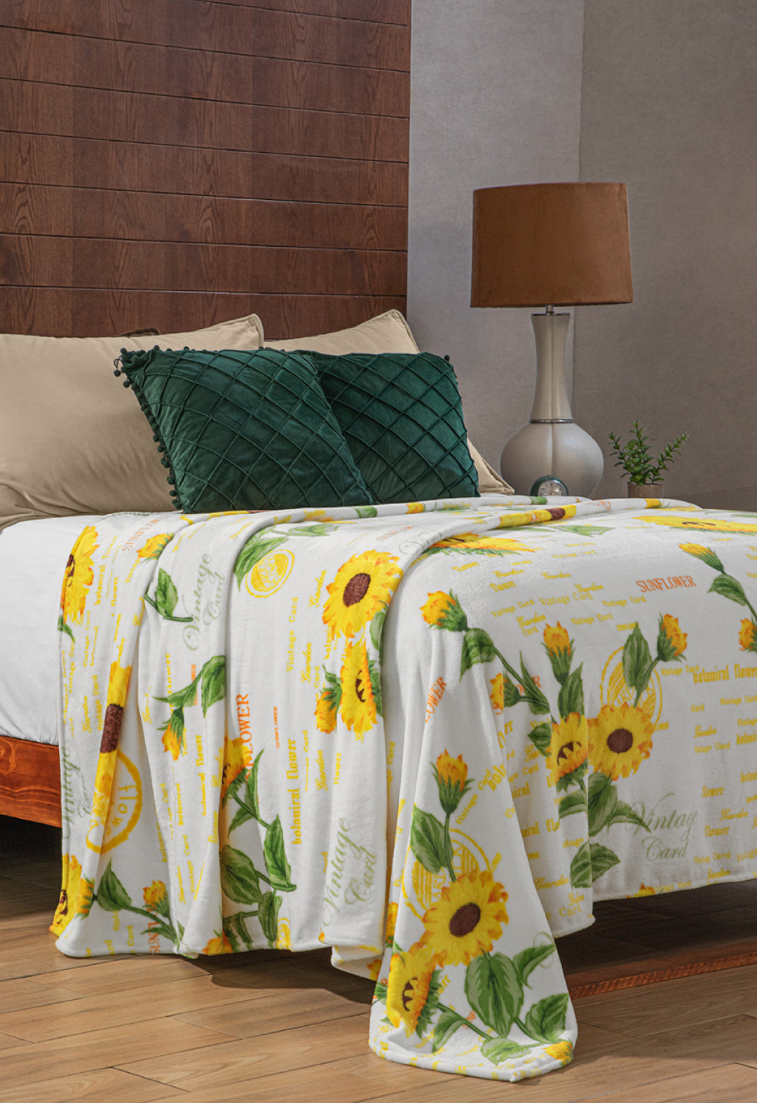 A cozy bed with beige pillows, a dark green cushion, and the Cobertor Flannel Ligero Sun de Intima Hogar, featuring a botanical design with yellow sunflowers and green leaves, in a warm bedroom with modern lamp and wooden floor.