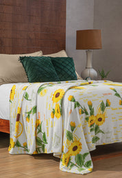 A cozy bed with beige pillows, a dark green cushion, and the Cobertor Flannel Ligero Sun de Intima Hogar, featuring a botanical design with yellow sunflowers and green leaves, in a warm bedroom with modern lamp and wooden floor.