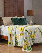 A neatly made bed with beige pillows, an Intima Hogar Cojín de Lujo Velvet Verde Esmeralda, a white sunflower-patterned blanket with yellow text, and dark green cushions. A brown lamp is on the bedside table in the wood-paneled room.