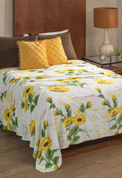 A neatly made bed features the Cobertor Flannel Ligero Sun by Intima Hogar with a bright yellow sunflower and green leaf design, two quilted yellow pillows, a brown headboard, and a nightstand with a brown lamp and small plant.