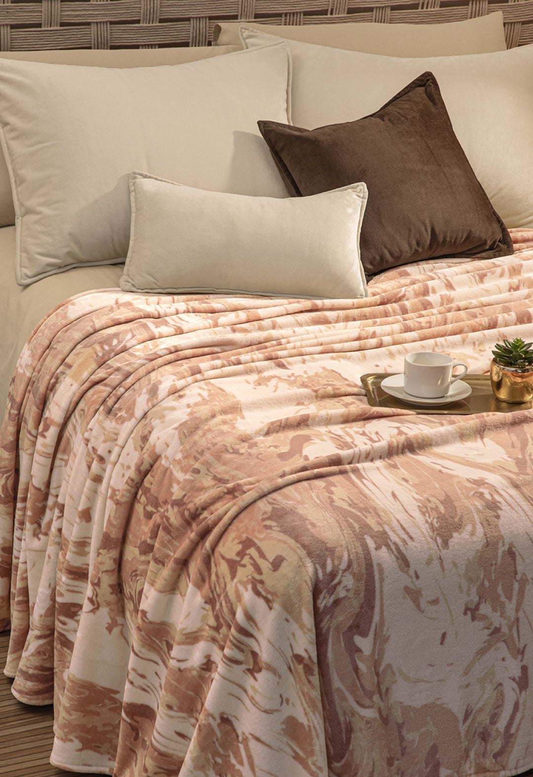 A neatly made bed with cream and brown pillows, an Intima Hogar Cobertor Flannel Ligero Tajín in marbled beige and pink, and a tray with a white cup, saucer, and a small potted plant.