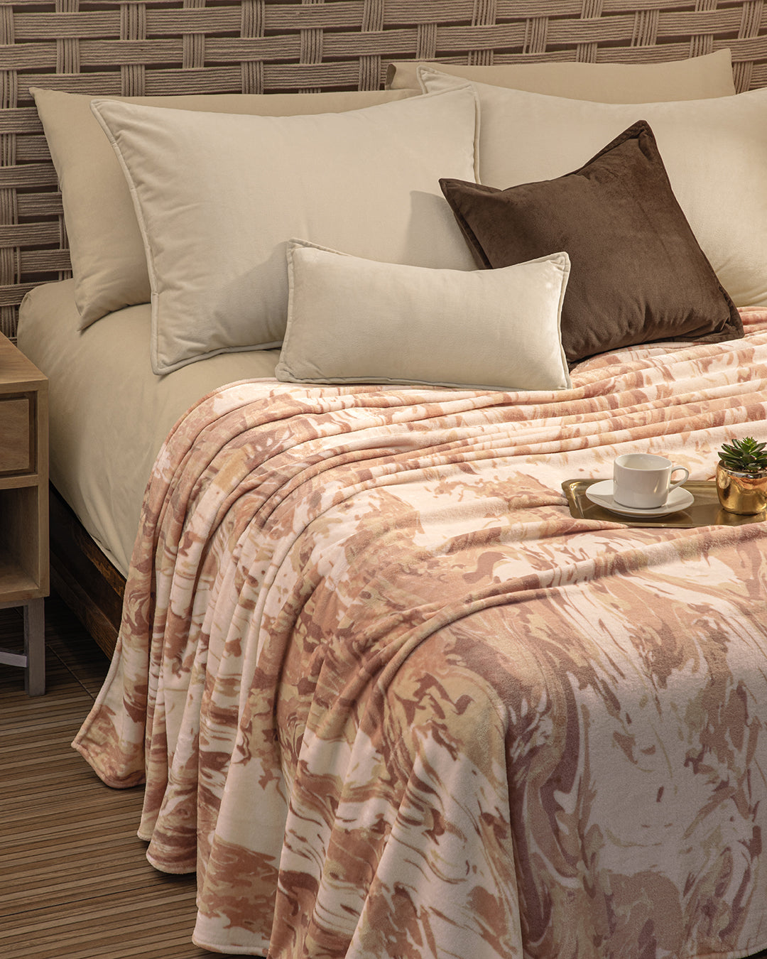 A neatly made bed features Intima Hogar’s Funda de Almohada Ensueño Beige, paired with brown pillows and a patterned pink and beige blanket. A tray with a white cup sits atop, while a small plant decorates the bedside next to the woven headboard.