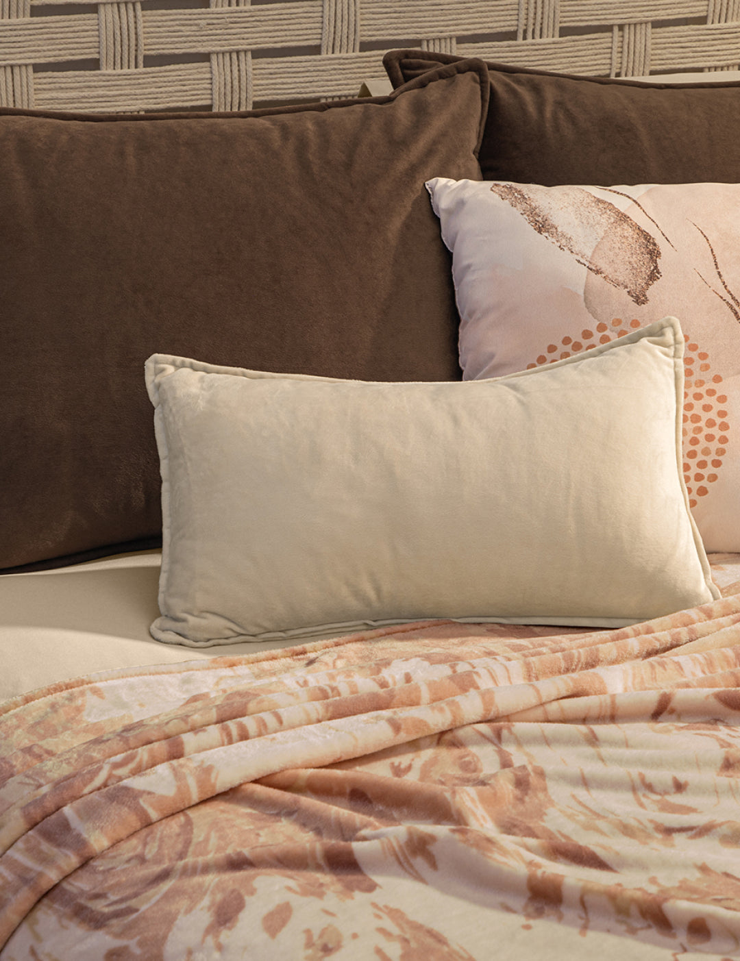 A cream pillow and a brown pillow rest against a textured headboard, with the Intima Hogar Cobertor Flannel Ligero Tajín in light pink and brown draped over the bed.
