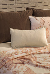 A cream pillow and a brown pillow rest against a textured headboard, with the Intima Hogar Cobertor Flannel Ligero Tajín in light pink and brown draped over the bed.