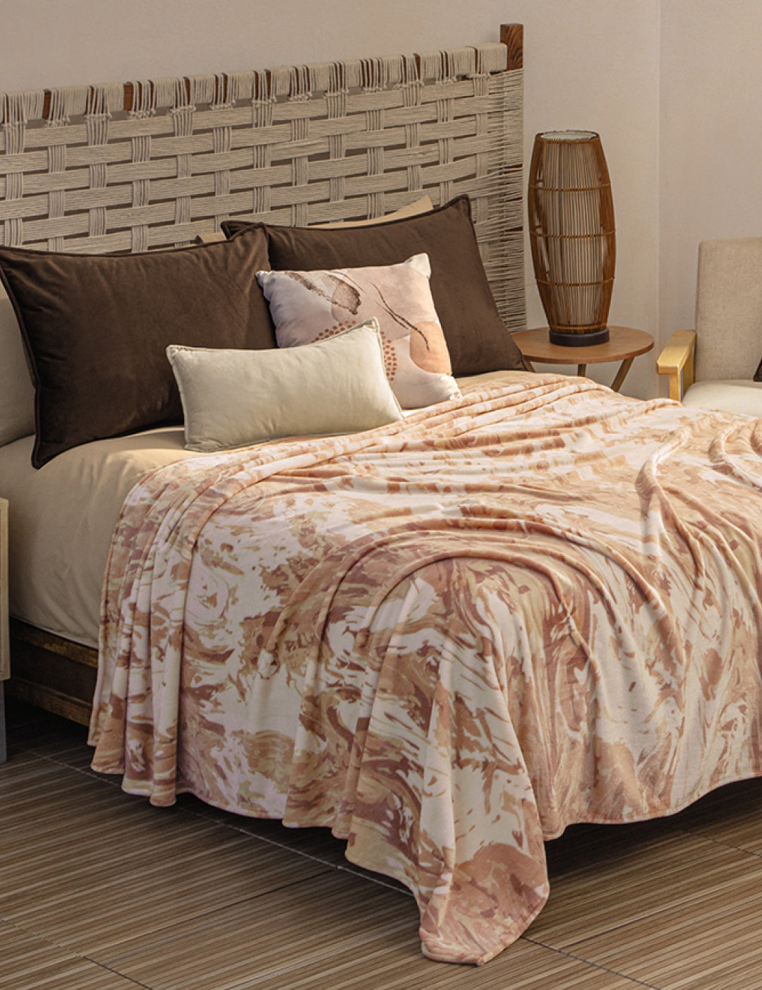 A cozy bed with a woven headboard, dark and light pillows, and the Cobertor Flannel Ligero Tajín by Intima Hogar in beige and pink. A round wooden side table with a tall lamp and a light armchair sit in a softly lit room.