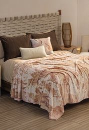 A cozy bed with a woven headboard, dark and light pillows, and the Cobertor Flannel Ligero Tajín by Intima Hogar in beige and pink. A round wooden side table with a tall lamp and a light armchair sit in a softly lit room.