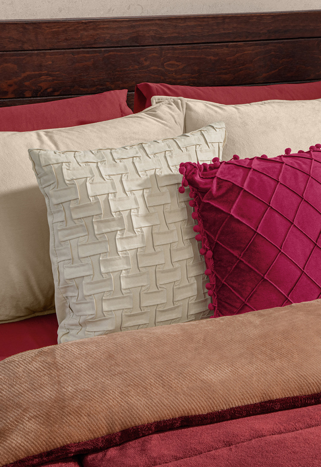 A neatly made bed with cream and red patterned pillows, layered with the Intima Hogar Cobertor Flannel Extra Suave Viñedo—perfect for cozy invierno nights—set against beige pillows and a wooden headboard.