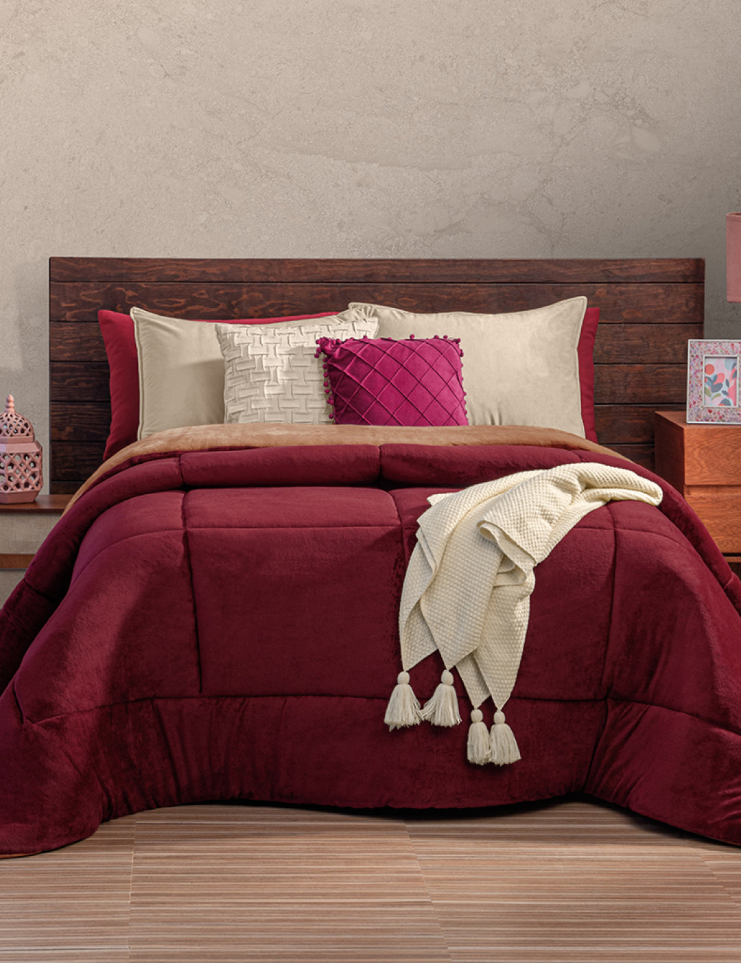 A neatly made bed with a dark wooden headboard, burgundy comforter, beige and burgundy pillows, and a cream knit throw—perfect for invierno with the Cobertor Flannel Extra Suave Viñedo by Intima Hogar for cozy warmth.