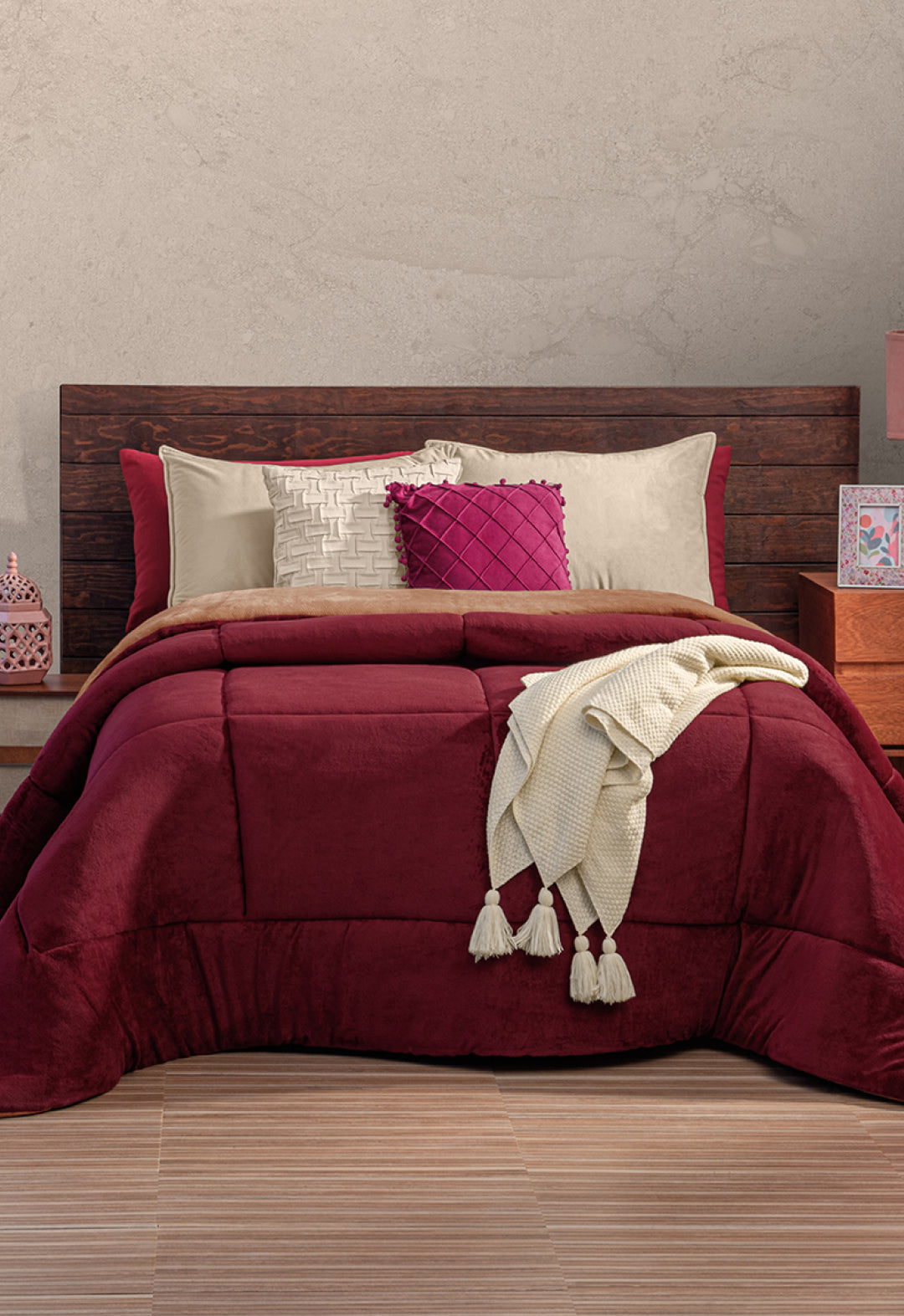 A neatly made bed with a dark wooden headboard, burgundy comforter, beige and burgundy pillows, and a cream knit throw—perfect for invierno with the Cobertor Flannel Extra Suave Viñedo by Intima Hogar for cozy warmth.