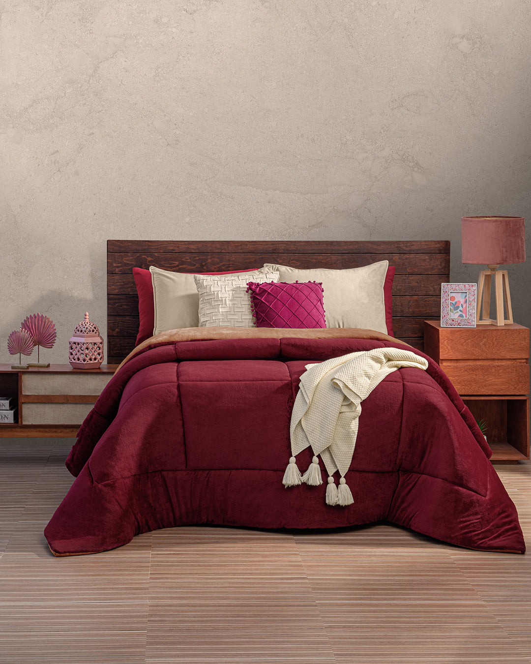 A neatly made bed features the Cojín de Lujo Velvet Vino by Intima Hogar, cream and pink pillows, and a cream throw. The wooden headboard sits against a light wall, flanked by nightstands with lamps, decor, and framed photos.