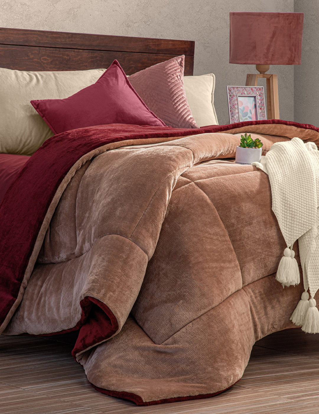 A cozy bed with the Intima Hogar Cobertor Flannel Extra Suave Viñedo in brown and burgundy, beige sheets, matching pillows, a white knit throw with tassels, a potted plant, and a lamp on a wooden nightstand—perfect for invierno.