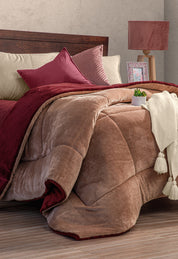 A cozy bed with the Intima Hogar Cobertor Flannel Extra Suave Viñedo in brown and burgundy, beige sheets, matching pillows, a white knit throw with tassels, a potted plant, and a lamp on a wooden nightstand—perfect for invierno.