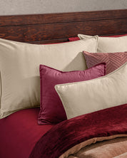 La cama luce la Funda de Almohada Ensueño Beige de Intima Hogar, manta burdeos y cabecero de madera marrón. Los cojines grandes van detrás y los pequeños al frente, creando un ambiente acogedor.