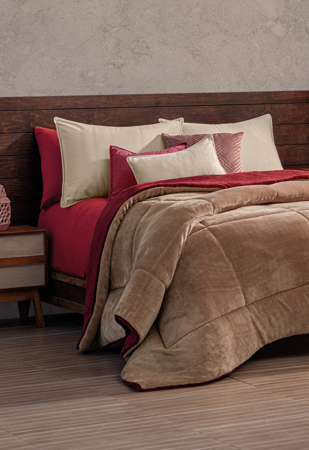 A neatly made bed features the Intima Hogar Cobertor Flannel Extra Suave Viñedo in beige and burgundy with matching pillows, a wood headboard, and nightstand with a pink decor piece. The wood floor and textured gray wall create a cozy invierno vibe.
