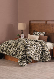 A neatly made bed with a brown wooden headboard features the Cobertor Platinum Extra Suave Finlandia by Intima Hogar in camuflaje verde musgo, complemented by several beige and brown pillows. A nightstand with a lamp, plant, and photo frame sits beside it on a tiled floor.