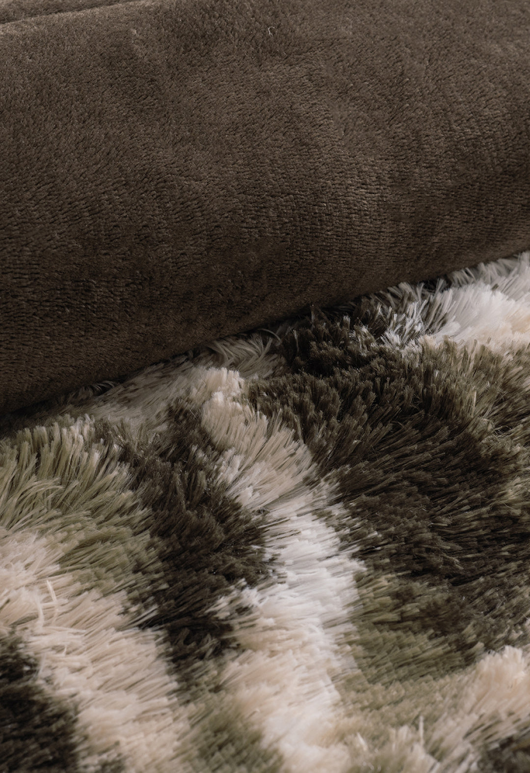 A close-up of the Cobertor Platinum Extra Suave Finlandia by Intima Hogar, rolled up on a shaggy, patterned carpet in moss green camo, brown, and white tones.