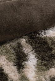A close-up of the Cobertor Platinum Extra Suave Finlandia by Intima Hogar, rolled up on a shaggy, patterned carpet in moss green camo, brown, and white tones.