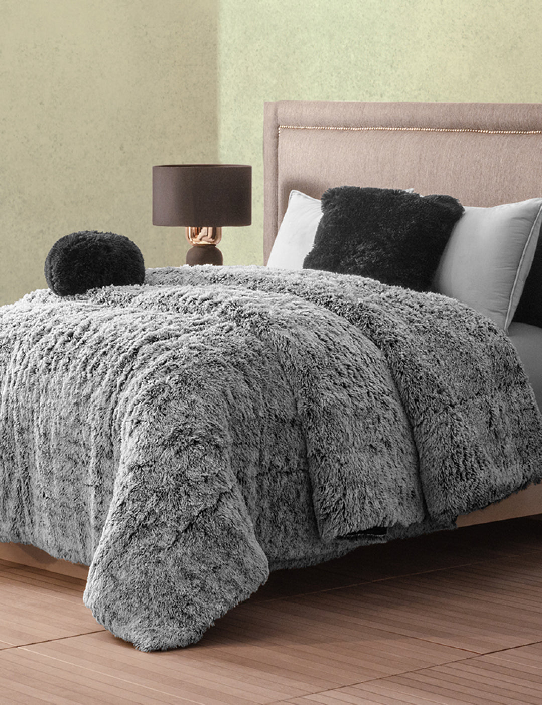 A cozy bedroom features a bed with the Cobertor Platinum Extra Suave Humo by Intima Hogar, matching pillows, and a round cushion. A lamp with a dark base and brown shade sits on the nightstand beside the upholstered headboard.