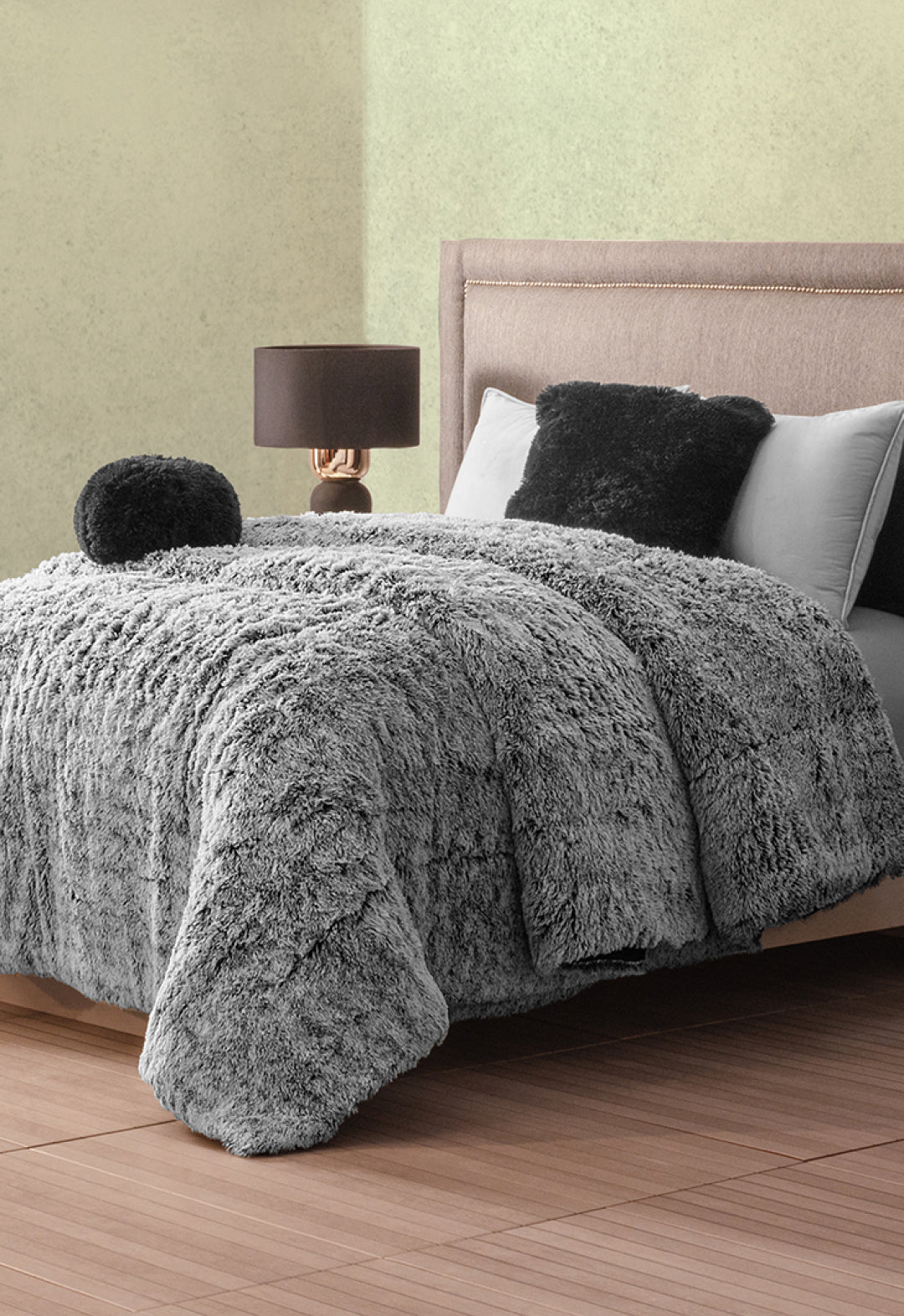 A cozy bedroom features a bed with the Cobertor Platinum Extra Suave Humo by Intima Hogar, matching pillows, and a round cushion. A lamp with a dark base and brown shade sits on the nightstand beside the upholstered headboard.