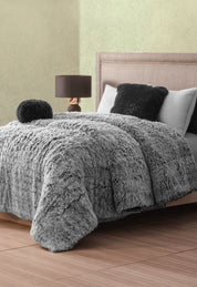 A cozy bedroom features a bed with the Cobertor Platinum Extra Suave Humo by Intima Hogar, matching pillows, and a round cushion. A lamp with a dark base and brown shade sits on the nightstand beside the upholstered headboard.