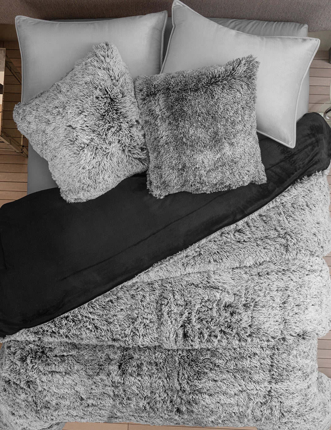 A neatly made bed with two gray pillows, two gray shaggy throw pillows, a black blanket, and a soft Cobertor Platinum Extra Suave Humo by Intima Hogar, all set on a wooden floor.