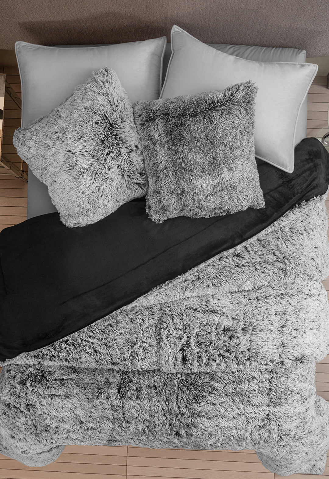 A neatly made bed with two gray pillows, two gray shaggy throw pillows, a black blanket, and a soft Cobertor Platinum Extra Suave Humo by Intima Hogar, all set on a wooden floor.