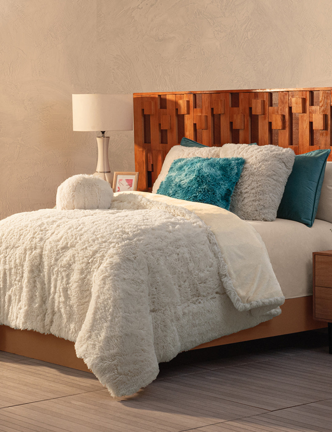 A cozy bedroom with a textured wooden headboard, a bed topped with the Cobertor Platinum Extra Suave Iglú by Intima Hogar, and franela pillows in white, teal, and turquoise. A white lamp and framed picture bring warmth to the bedside table.