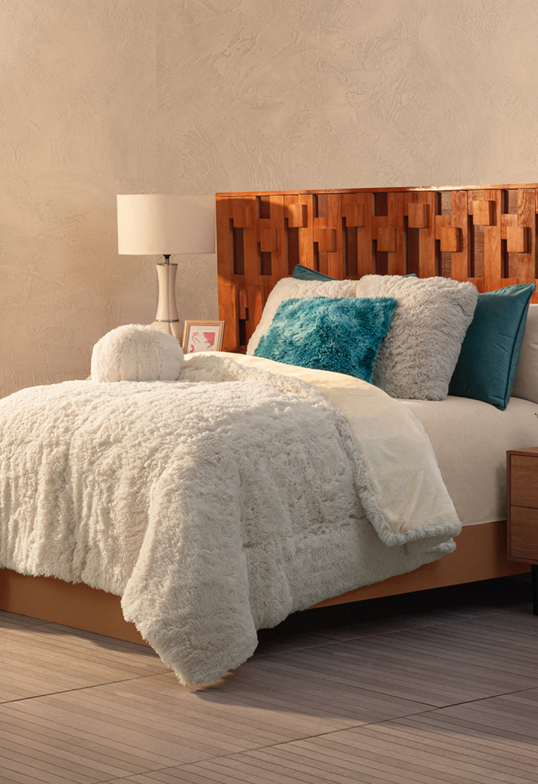 A cozy bedroom with a textured wooden headboard, a bed topped with the Cobertor Platinum Extra Suave Iglú by Intima Hogar, and franela pillows in white, teal, and turquoise. A white lamp and framed picture bring warmth to the bedside table.