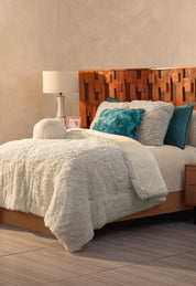 A cozy bedroom with a textured wooden headboard, a bed topped with the Cobertor Platinum Extra Suave Iglú by Intima Hogar, and franela pillows in white, teal, and turquoise. A white lamp and framed picture bring warmth to the bedside table.