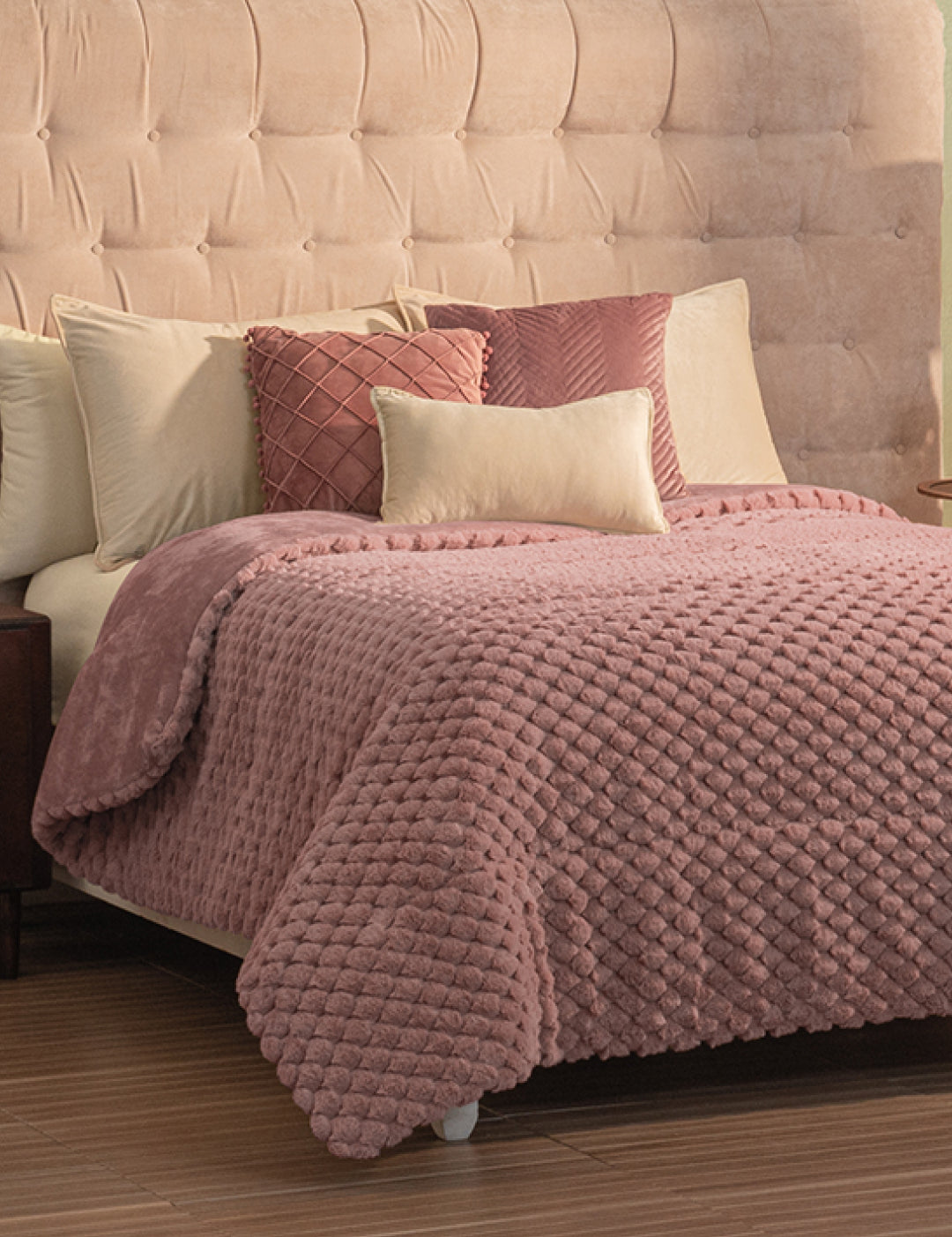 A neatly made bed with a beige tufted headboard, beige and pink pillows, and a textured pink Cobertor Platinum Extra Suave Aruba by Intima Hogar, set on a wooden floor.