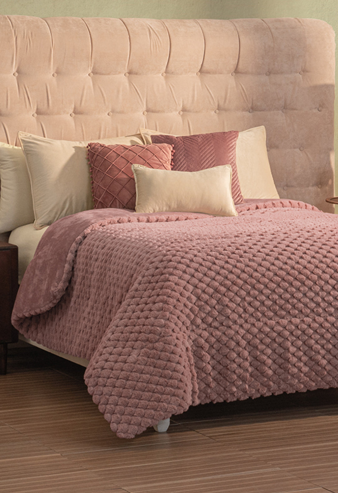 A neatly made bed with a beige tufted headboard, beige and pink pillows, and a textured pink Cobertor Platinum Extra Suave Aruba by Intima Hogar, set on a wooden floor.