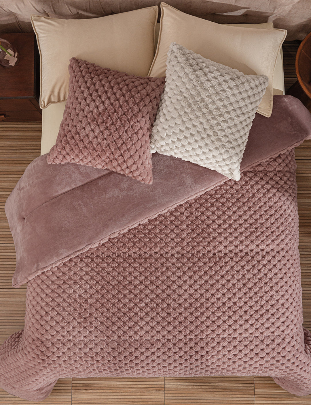 A neatly made bed features the Cobertor Platinum Extra Suave Aruba by Intima Hogar in mauve, with matching mauve and off-white textured pillows, beige sheets and pillows, all set on a light wood floor beside a wooden nightstand.