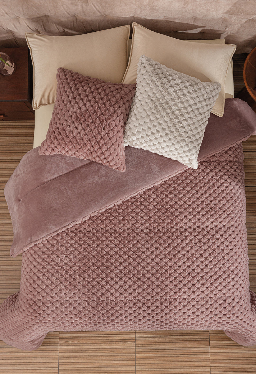 A neatly made bed features the Cobertor Platinum Extra Suave Aruba by Intima Hogar in mauve, with matching mauve and off-white textured pillows, beige sheets and pillows, all set on a light wood floor beside a wooden nightstand.
