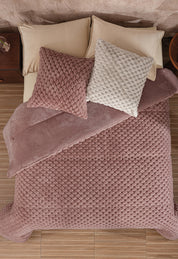 A neatly made bed features the Cobertor Platinum Extra Suave Aruba by Intima Hogar in mauve, with matching mauve and off-white textured pillows, beige sheets and pillows, all set on a light wood floor beside a wooden nightstand.