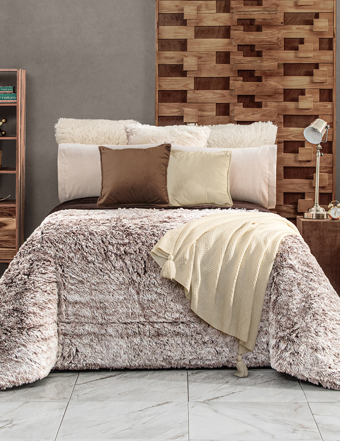A cozy bed with a fluffy Cobertor Platinum Extra Suave Glacial by Intima Hogar in beige, three pillows in varied tones, a cream knit throw, and a wooden accent wall. A lamp and books rest on bedside tables over a white marble floor.