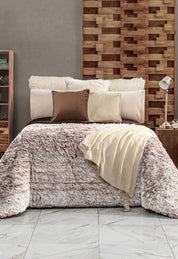 A cozy bed with a fluffy Cobertor Platinum Extra Suave Glacial by Intima Hogar in beige, three pillows in varied tones, a cream knit throw, and a wooden accent wall. A lamp and books rest on bedside tables over a white marble floor.