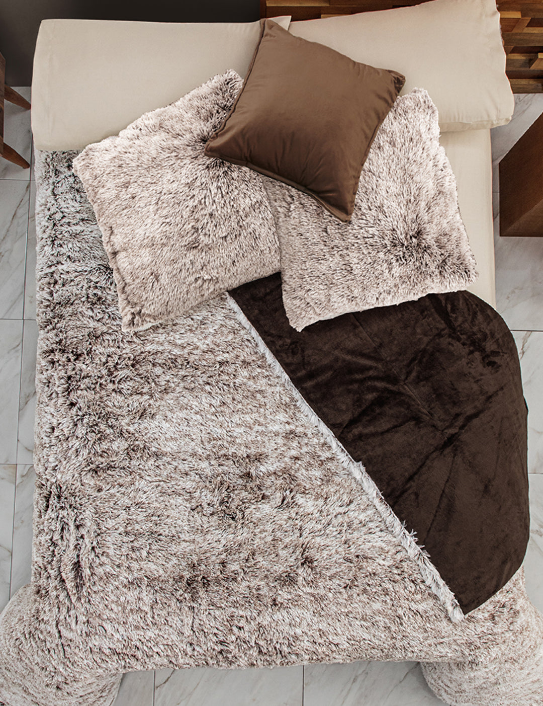 A bed with cream sheets, two large beige pillows, a small brown satin pillow, and an Intima Hogar Cobertor Platinum Extra Suave Glacial in beige and brown, partially folded back, arranged on a white tiled floor.