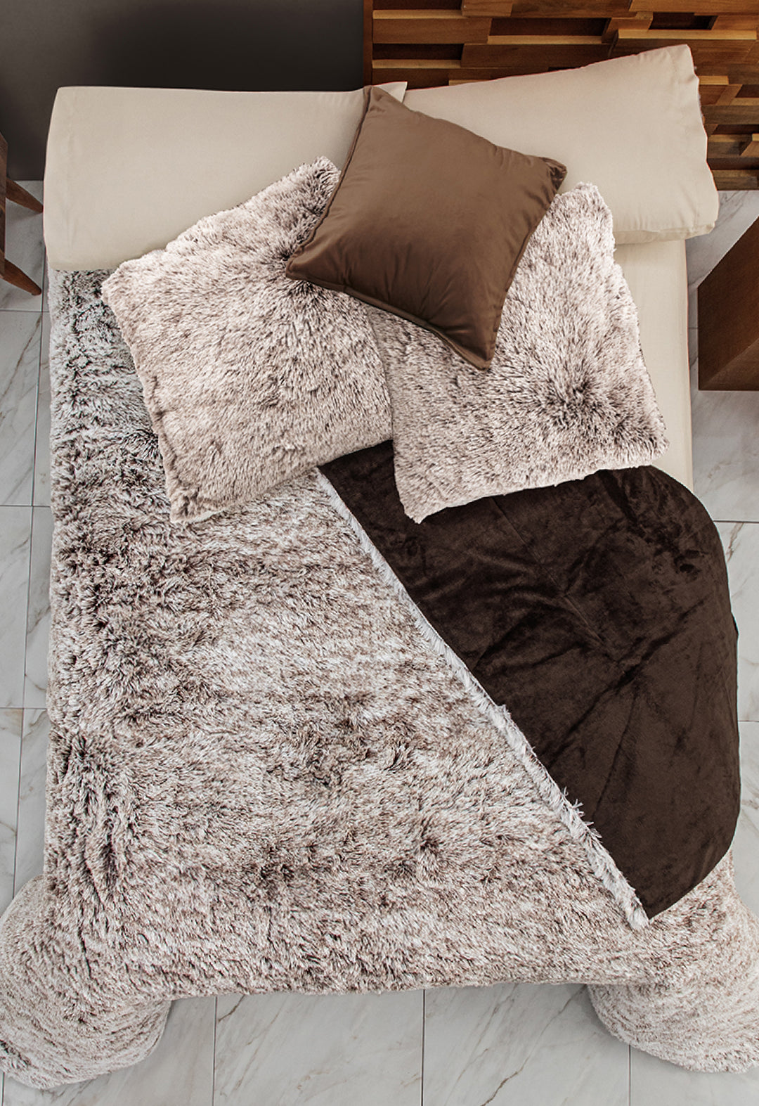 A bed with cream sheets, two large beige pillows, a small brown satin pillow, and an Intima Hogar Cobertor Platinum Extra Suave Glacial in beige and brown, partially folded back, arranged on a white tiled floor.
