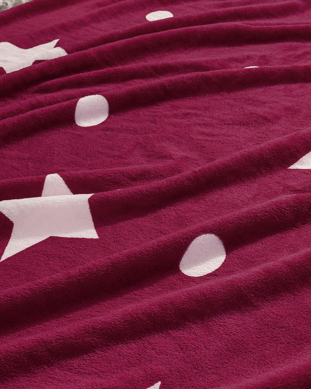 A close-up of the Frazada Saturno by Intima Hogar in maroon, adorned with white stars and circles. This plush, velvety blanket features gentle folds, blending warmth and style for your home.