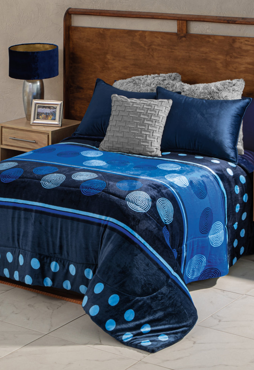 A neatly made bed with the Cobertor Flannel Extra Suave Sfera by Intima Hogar, blue and gray pillows, and a wooden headboard creates a stylish look. A nightstand with a blue lamp and framed photo adds a modern touch for recámaras modernas.